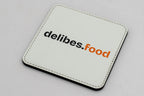 Quadratischer weißer Untersetzer mit schwarzem und orangefarbenem delibes.Food-Logo.