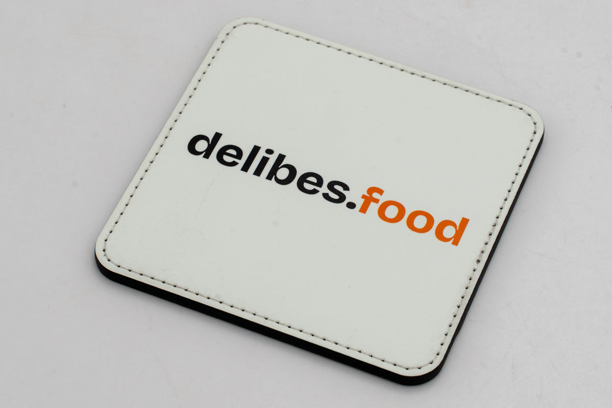 Quadratischer weißer Untersetzer mit schwarzem und orangefarbenem delibes.Food-Logo.