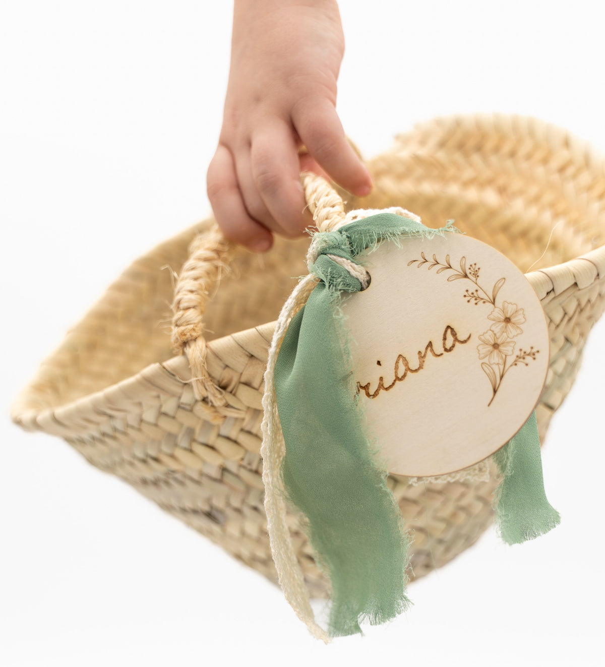 Mano infantil sosteniendo un cesto de palma con lazo verde y etiqueta de madera con nombre.