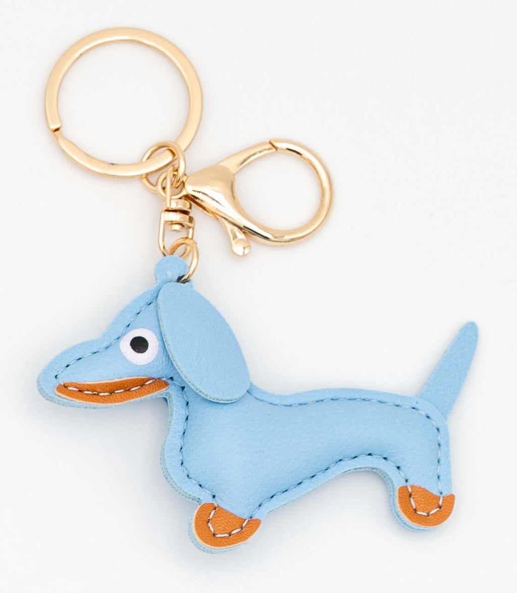 Colgante de perro salchicha en polipiel azul con herrajes dorados y costuras blancas.