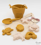 Cubo de playa color mostaza personalizado con nombre láser y bolsa de malla beige.