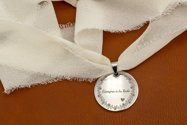 Anhänger aus spiegelpoliertem Edelstahl mit dem Spruch „Immer an deiner Seite“ und Blumenrand, an einem elfenbeinfarbenen Chiffonband